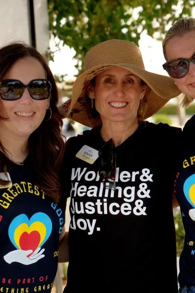 Three women smiling in black T-shirts. Two of the shirts say: The Greater Good. The center shirt says: Wonder& Healing& justice& joy.