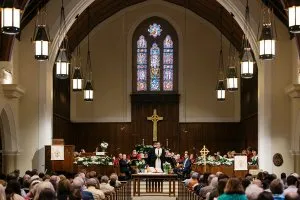 People sit in pews looking toward a man in black robes and a white stole who stands at a table in front of a carved cross and a stained glass window.