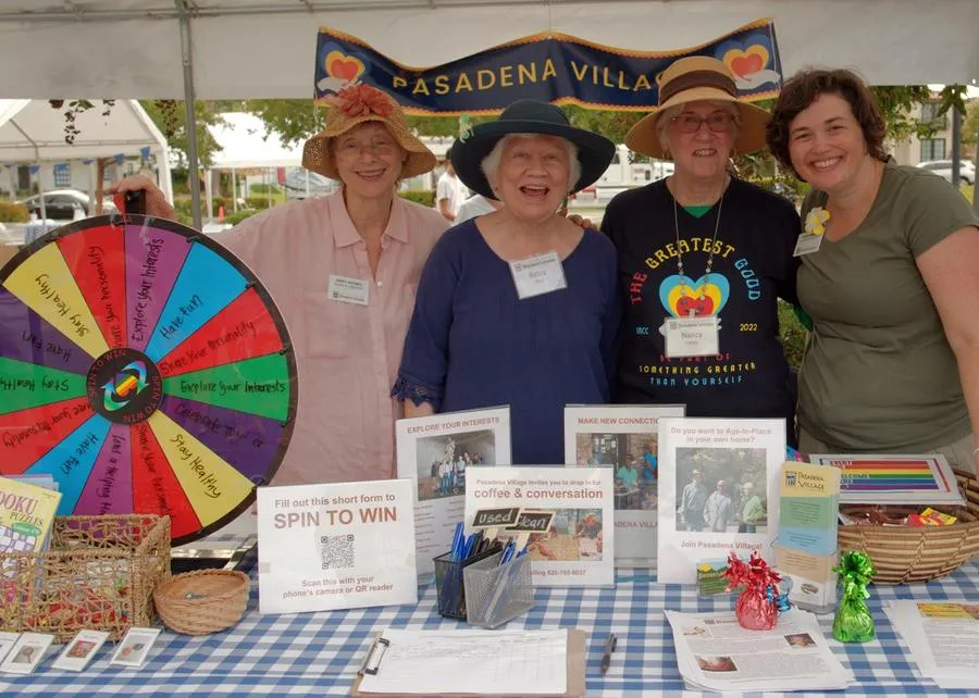 Four women stand behind a gingham covered table with a prize wheel.