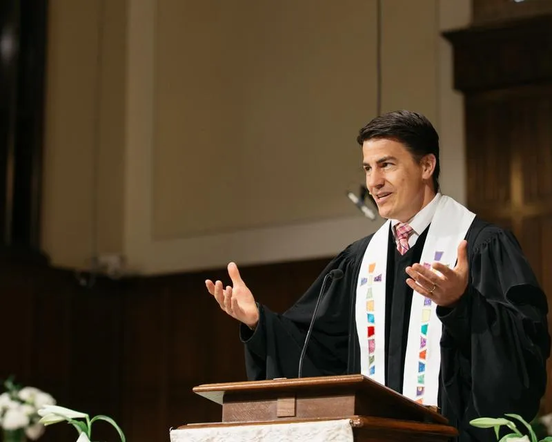 Rev. Dr. Erik Wiebe preaching from the pulpit while wearing black robes and a multi-colored stole.