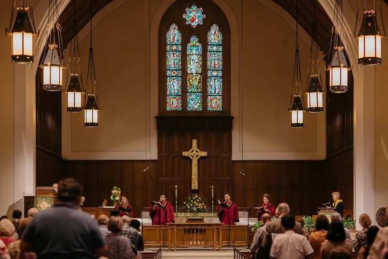 Four people in red robes sing in the wood paneled front apse of SMCC. A carved cross hangs under three stained glass windows behind them.