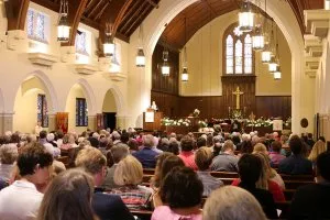 The SMCC sanctuary with white plaster walls, white stone side colonnades, and a wood panelled front apse and arched ceiling. A woman preaches from the dark wood pulpit at the front left of the apse. The pews are filled with people.