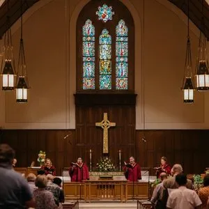Four singers in red robes sing in the front apse of the SMCC sanctuary. There is an altar, cross, and stained glass window behind them.