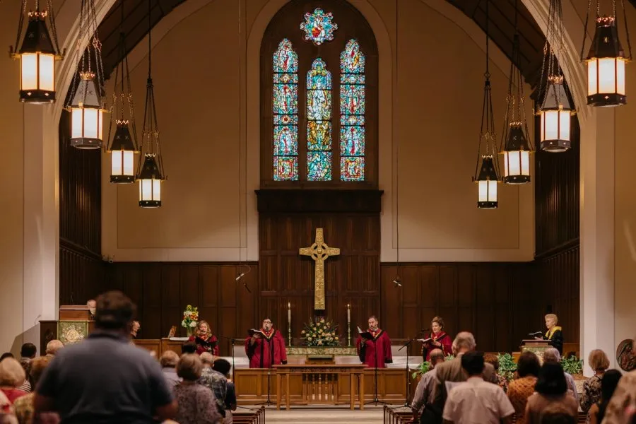 Four singers in red robes sing in the front apse of the SMCC sanctuary. There is an altar, cross, and stained glass window behind them.