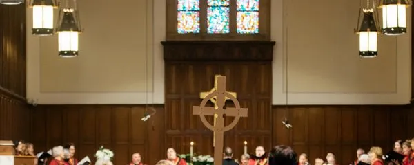 The backs of three people in black vestments and white stoles following a cross down a center aisle past people sitting in pews inside the SMCC sanctuary.