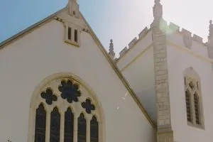 The west facade of SMCC. Five narrow stained glass windows are crowed by three rose windows under a pointed roof and a stone cross, next to the square bell tower.